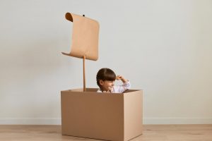 portrait of charming little girl sitting in cardboard box images she is sailing boat looking far exploring horizon keeping hand above eyes with happy toothy smile juegos reciclaje
