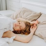 Una chica joven de pelo rizado y rubio durmiendo plácidamente y relajada en una cama cómoda con edredón blanco y almohadas beige, representando el descanso profundo y los sueños con dinero o prosperidad.